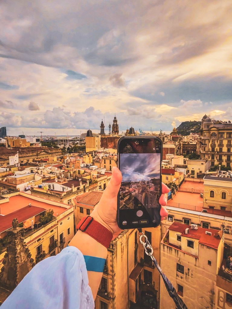 A hand holds a smartphone to photograph Barcelona rooftops beneath a cloudy sky.
