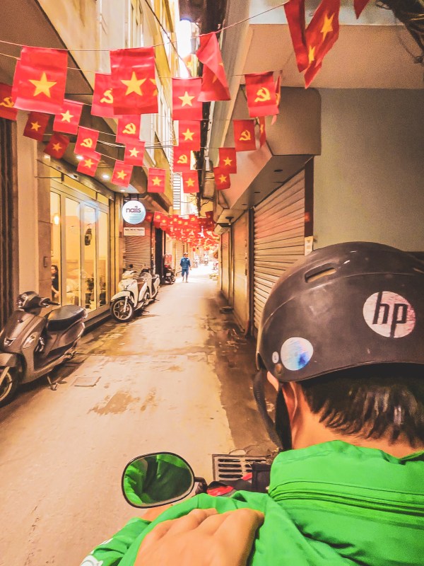 A rider in a green helmet travelling through a flag-lined Hanoi lane.