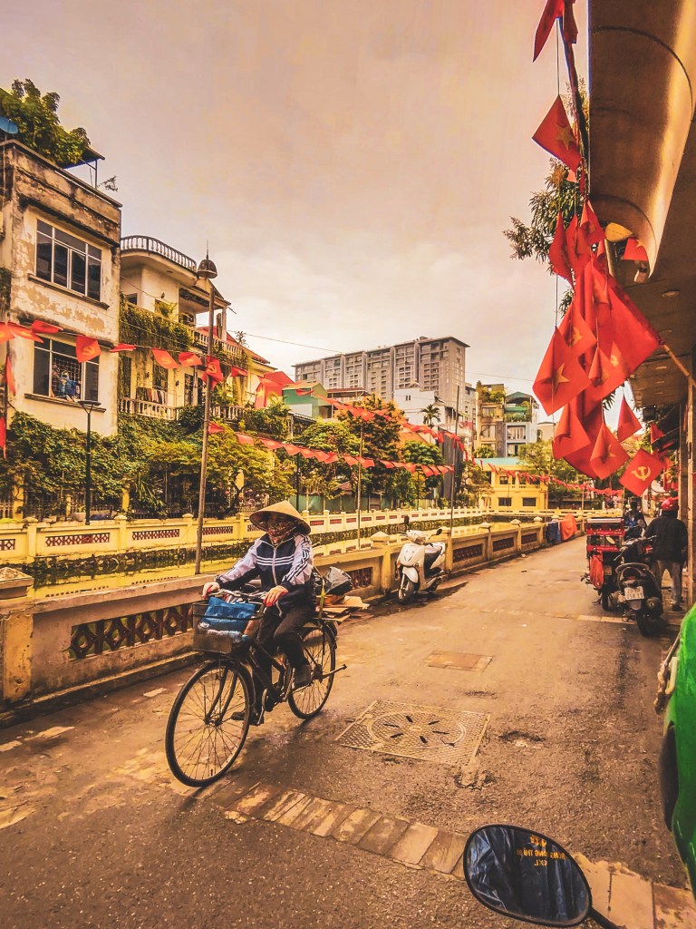 A cyclist rides along a Hanoi street beside hanging flags and flowers.