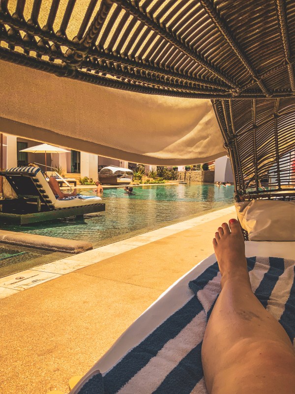 A guest reclining beside a shaded pool under a slatted roof.