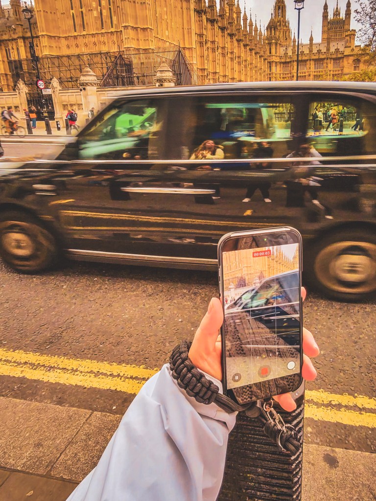 A smartphone recording a passing black taxi outside the Palace of Westminster in London.