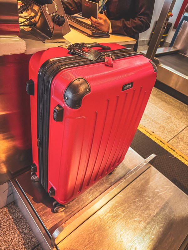 A red hard-shell suitcase with luggage visible behind it.