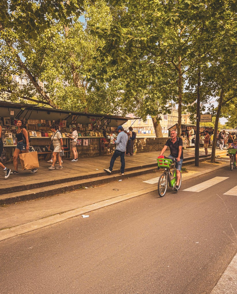 A cyclist passes tree-shaded bookstalls along the Seine in Paris.