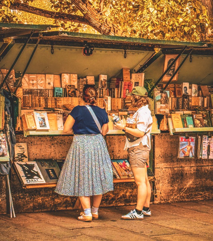 Two people browsing books at an outdoor bookstall beneath leafy trees in Paris.