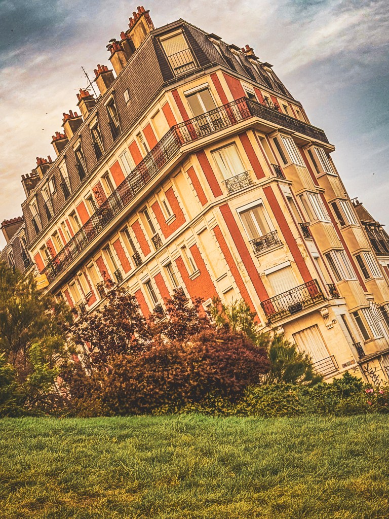 A red-and-cream Paris corner building above a grassy slope and flowering shrubs.