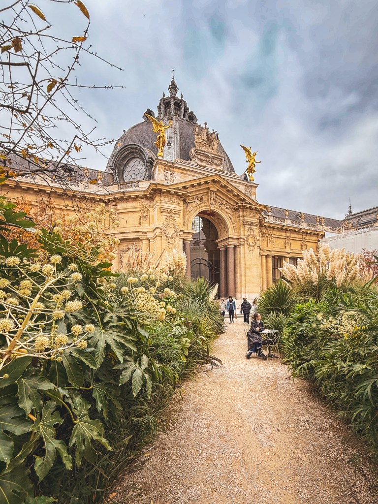 A garden path framed by plants leads toward an ornate building in Paris.