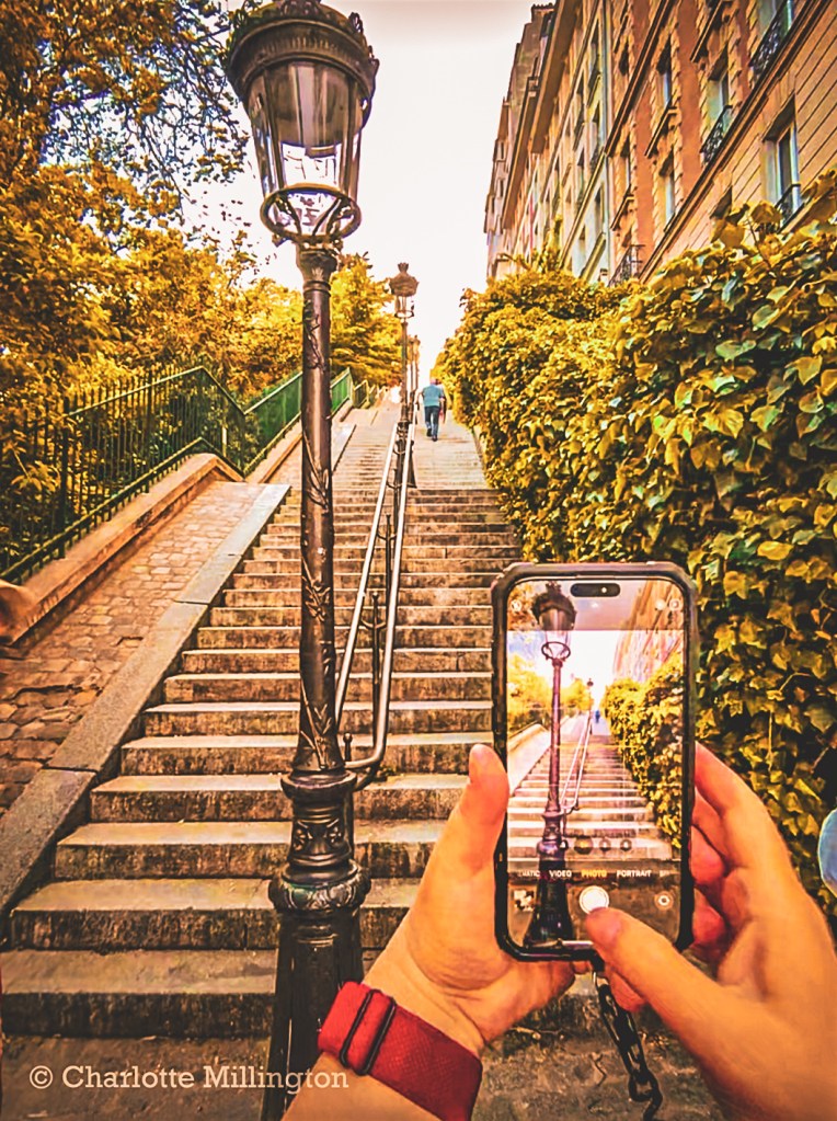 Hands holding a smartphone to photograph a tree-lined staircase and streetlamp in Paris.