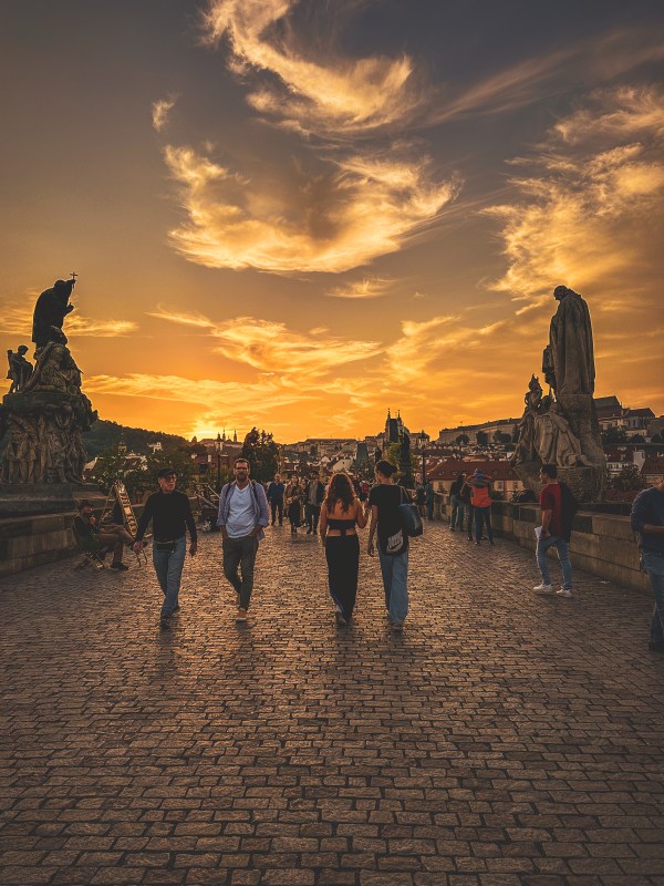 People walking among bridge statues beneath a sunset in Prague.