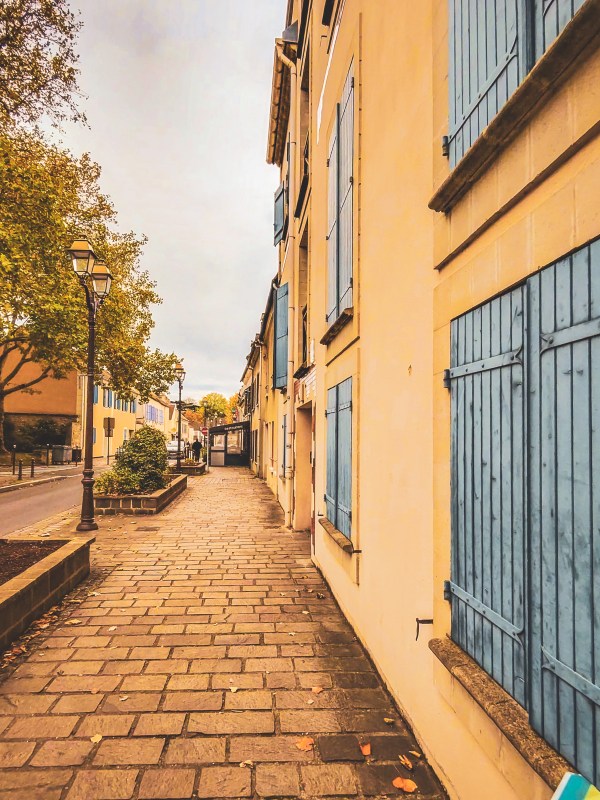 A paved lane beside yellow walls and blue shutters in Roissy-en-France.