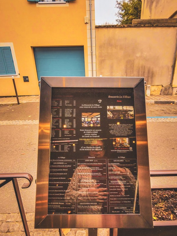 An outdoor menu board beside a yellow building in Roissy-en-France.