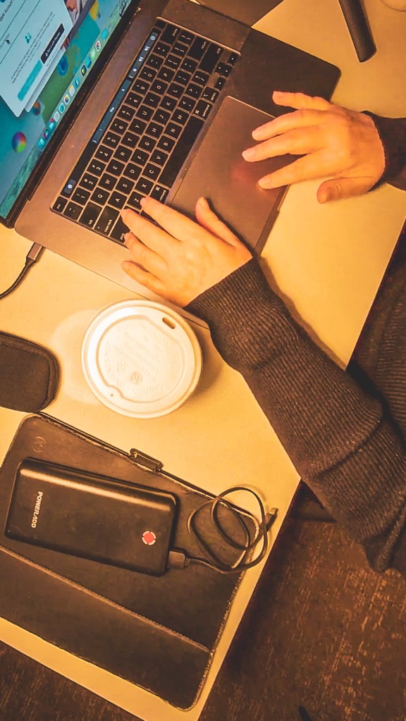 Hands working at a laptop beside a notebook, portable drive and coffee.