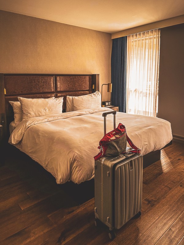 Hotel bed beside a suitcase and backpack.